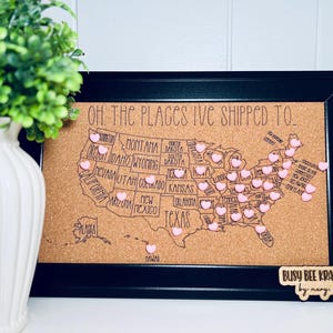 May include: A corkboard map of the United States with pink heart-shaped push pins marking locations. The map is framed in black and has the text "OH. THE PLACES I'VE SHIPPED TO" at the top. The bottom right corner has a small wooden sign that reads "BUSY BEE KRAFTS by nancy.llc".
