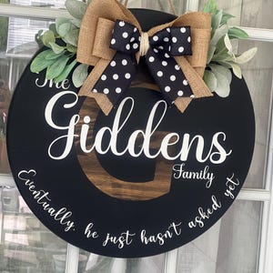 May include: A black round wooden door hanger with the text "The Giddens Family Eventually he just hasn't asked yet". The door hanger is decorated with a burlap and black and white polka dot bow and greenery.