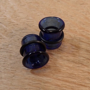 May include: Two dark blue glass ear plugs with black rubber O-rings. The plugs have a flared design and are sitting on a light brown wooden surface. The plugs are designed for stretched ear piercings.