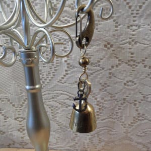 May include: A gold-toned bell charm with an anchor pendant. The charm is attached to a silver-toned safety pin.