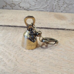 May include: A small brass bell with an anchor charm and a clasp. The bell has a decorative pattern on the side.