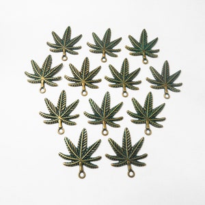May include: An overhead shot displays eleven bronze-colored cannabis leaf charms. Each charm has a detailed leaf design with visible veins and a small loop at the bottom for attachment. The charms are arranged in a loose, symmetrical pattern against a white background.