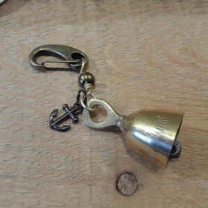 May include: A gold-colored bell charm with an anchor charm and a black bead. The bell has a small loop at the top and a clip on the other end.