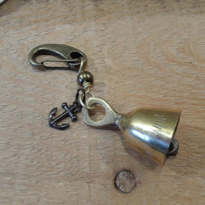 May include: A gold-colored bell charm with an anchor charm and a black bead. The bell has a small loop at the top and a clip on the other end.