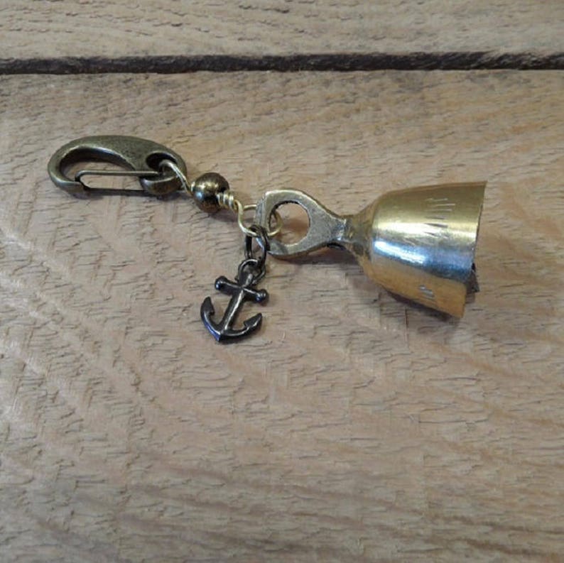 May include: A gold-toned bell charm with an anchor charm attached. The bell has a small loop at the top and a clip on the other end.