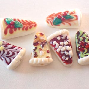 Op de afbeelding: Zes decoratieve glazen kralen in de vorm van taartpunten. De ontwerpen omvatten rode en witte wervelingen, bessen en groene bladeren, met een witte glazuurbasis. Perfect voor sieraden maken.