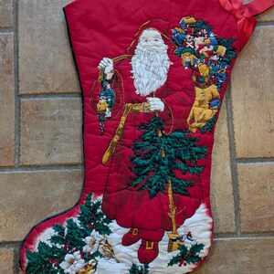 May include: Red Christmas stocking with a quilted design featuring Santa Claus. The stocking depicts Santa holding a lantern and a Christmas tree. The stocking is decorated with images of toys, birds, and holly. A red ribbon is attached.