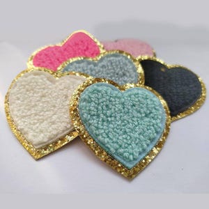 Könnte beinhalten: Eine Sammlung herzförmiger Aufnäher in verschiedenen Farben, darunter Rosa, Weiß, Türkis und Grau. Jeder Aufnäher hat eine strukturierte, flauschige Oberfläche und ist mit goldenem Glitzer umrandet. Diese Aufnäher eignen sich ideal zum Basteln und Verzieren von Kleidung oder Accessoires.