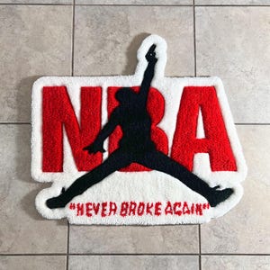 Könnte beinhalten: Ein getufteter Teppich mit weißem Hintergrund und großen roten Buchstaben, die NBA bilden. Eine schwarze Silhouette einer springenden Figur ist über den Buchstaben zentriert. Unter der Figur steht der Text NEVER BROKE AGAIN in roten Großbuchstaben.