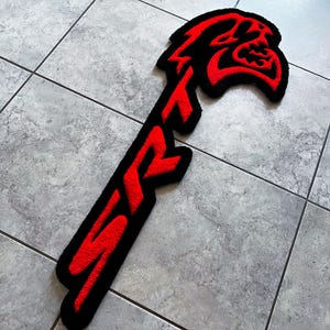 May include: A black and red rug in the shape of a stylized logo. The logo features the letters "SRT" in red, with a red and black graphic of a snarling face at the top. The rug is placed on a tiled floor.