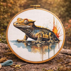 Peut inclure: Broderie au point de croix d'un gecko dans un cercle en bois. Le gecko est représenté dans des tons de jaune, de marron et de bleu, avec une touche de peinture colorée. Le cercle à broder repose sur une pierre, avec du fil et des ciseaux à proximité.
