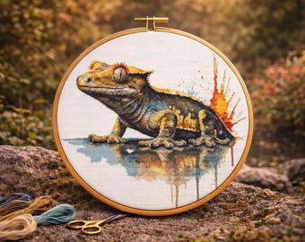 Könnte beinhalten: Kreuzstich-Stickerei eines Geckos in einem Holzreifen. Der Gecko ist in Gelb-, Braun- und Blautönen dargestellt, mit einem farbenfrohen Farbspritzer. Der Stickrahmen liegt auf einem Stein, mit Faden und Schere in der Nähe.