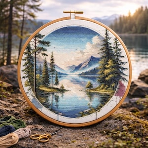 May include: A completed cross-stitch landscape scene of a lake surrounded by evergreen trees and mountains, set in a wooden embroidery hoop. The scene is detailed with blue, green, and brown threads. Embroidery floss and scissors are in the foreground.