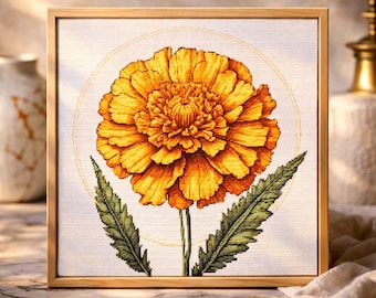May include: A framed cross-stitch depicting a marigold flower. The flower is a bright orange with detailed petals, a green stem, and two green leaves. The artwork is set against a white background and framed in light wood.