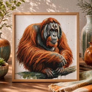 May include: A framed cross-stitch of an orangutan. The artwork features the primate with reddish-brown fur, gray face, and is set against a white background. The orangutan is sitting on a branch. The frame is made of light-colored wood.