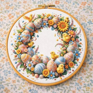 May include: A cross-stitch embroidery hoop with an Easter wreath design. The wreath includes decorated Easter eggs, yellow chicks, and colourful flowers. The embroidery is set against a white fabric background, framed by a wooden hoop.