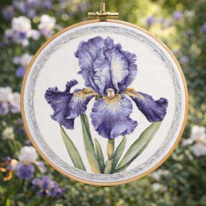 Può includere: Un ricamo a punto croce di un fiore di iris viola in un telaio di legno. Il fiore ha petali dettagliati con accenti bianchi e gialli, foglie verdi e un bordo decorativo. Lo sfondo è sfocato con altri fiori.
