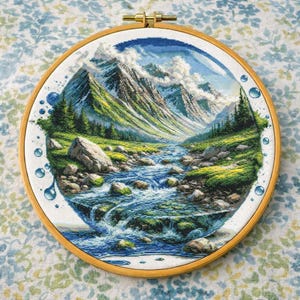 Puede incluir: Bordado de punto de cruz de un paisaje de montaña, enmarcado en un aro de madera redondo. La escena representa un río que fluye por un valle, con montañas nevadas al fondo. El bordado utiliza tonos de azul, verde y marrón.
