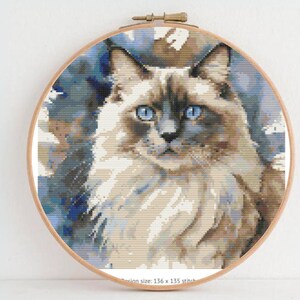 Peut inclure: Une broderie au point de croix d'un chat Ragdoll aux yeux bleus perçants, encadrée dans un cercle à broder en bois. Le pelage du chat est un mélange de beige, de marron et de gris, créant un aspect détaillé et texturé. La taille du motif est de 136 x 135 points.
