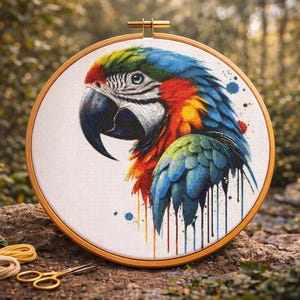 May include: A colorful parrot embroidery in a wooden hoop. The parrot features vibrant blue, green, red, and yellow feathers. The embroidery is set against a white background, with blue paint splatters. Embroidery floss and scissors are in the foreground.