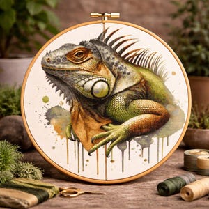 May include: A watercolor-style embroidery hoop featuring a detailed iguana illustration. The iguana is depicted in shades of green, brown, and orange, with a textured appearance. The artwork is set against a white background with paint splatters and a wooden hoop.