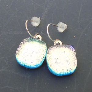 May include: A pair of dichroic glass earrings. Each earring features a rounded square shape with a silver hook. The glass has a white center with blue and green edges, creating a shimmering effect. The earrings are designed for pierced ears.