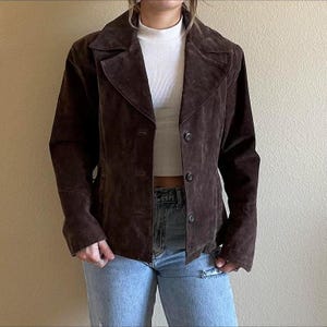 Puede incluir: Una chaqueta de ante marrón con cuello clásico y cierre de botones. La chaqueta tiene un corte entallado y mangas largas. La chaqueta se lleva sobre una camiseta blanca y unos vaqueros azules.