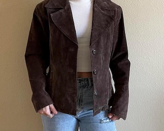 May include: A brown suede jacket with a classic collar and button closure. The jacket has a tailored fit and long sleeves. The jacket is worn over a white top and blue jeans.
