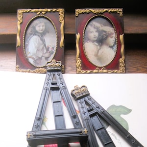 May include: Two antique oval portrait frames with gold trim and red backgrounds, each holding a sepia-toned photograph of a child. Two black miniature easels with gold accents and crown details are in the foreground, suggesting a vintage or decorative theme.