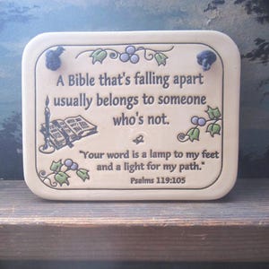 May include: A rectangular cream-colored ceramic plaque with rounded corners and two dark hanging cords. It features a printed illustration of an open book, a candle, and green leaves with purple berries, alongside the text: A Bible that's falling apart usually belongs to someone who's not. Your word is a lamp to my feet and a light for my path. Psalms 119:105.