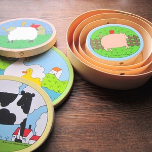 May include: Set of four wooden nesting boxes with colorful illustrations of farm animals. The boxes feature a sheep, a duck, a cow, and a pig. Each box has a different design and is decorated with bright colors.
