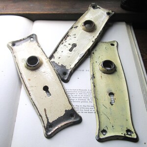 May include: Three rectangular metal door plates with a distressed cream-colored finish and visible dark metal underneath. Each plate features a central circular opening for a knob and a lower keyhole, with small screw holes at the top and bottom corners.