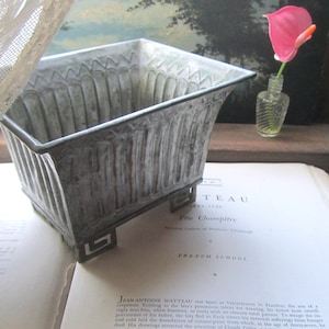 May include: A rectangular metal planter with a weathered gray finish and vertical ribbed detailing. The container features four small feet with a geometric cutout design. The exterior surface shows a distressed patina with darker gray accents.