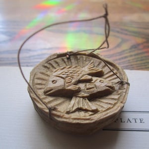 May include: A carved wooden ornament featuring two fish. The fish are intricately detailed and set within a circular frame. A thin string is attached for hanging. The ornament is a light brown color.