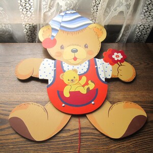 May include: A wooden pull toy of a brown bear wearing a blue and white striped hat and a red and blue overall with a yellow bear cub in the pocket. The bear is holding a red flower.