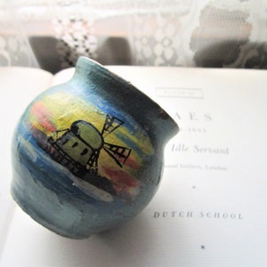 May include: A small, round, blue ceramic pot with a painted scene of a windmill and a sunset. The pot is hand-painted with yellow, orange, red, and green accents.