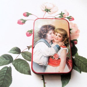 May include: A vintage tin box with a colorful image of two children, a boy and a girl, embracing. The boy is wearing a sailor shirt and the girl is wearing a white dress with a red sash. The tin box has a red border.