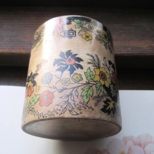 May include: A cylindrical container with a floral design. The container is beige with a pattern of colorful flowers in red, yellow, and blue. The flowers are set against a backdrop of black leaves and stems.