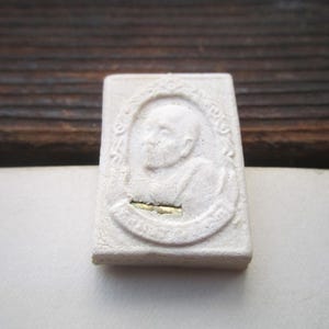 May include: White ceramic amulet with a raised image of a person's head and shoulders. The amulet is oval shaped and has a gold trim around the edge.