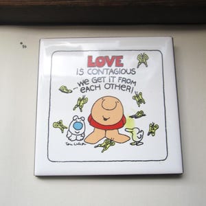 May include: Square ceramic tile with a cartoon illustration. The tile features a smiling, tan figure with a red collar, surrounded by butterflies and other cartoon characters. The text "LOVE is contagious, we get it from each other!" is written in red.
