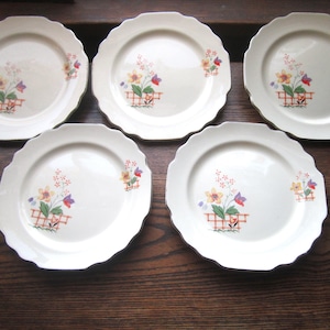May include: Set of five white ceramic plates with scalloped edges. Each plate features a floral design with yellow, orange, red, and purple flowers in a basket.