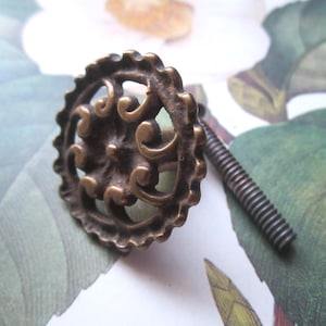 May include: A round, antique brass-toned metal cabinet knob featuring a scalloped edge and an intricate, openwork scroll pattern in the center. The knob includes a long, threaded metal bolt extending from the back for secure installation.