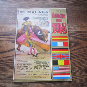May include: Vintage Spanish bullfighting poster from Malaga, featuring a matador in a colorful suit facing a bull. The poster has text in Spanish and includes flags of various countries. The poster is on a wooden surface.