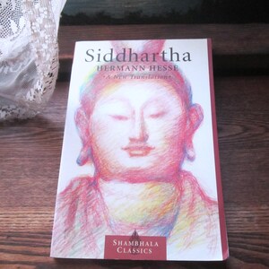 May include: A paperback book titled Siddhartha by Hermann Hesse, featuring a colorful, sketch-style illustration of a face on the white cover. The text reads Siddhartha, Hermann Hesse, A New Translation, and Shambhala Classics in a red box.