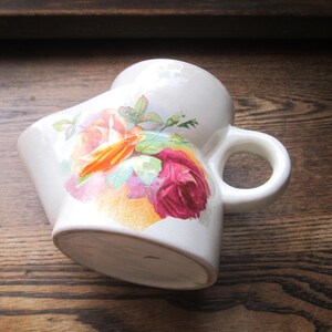May include: White ceramic shaving mug with a floral design featuring red, orange, and pink roses. The mug has a curved shape and a handle. The floral pattern is set against a white background.