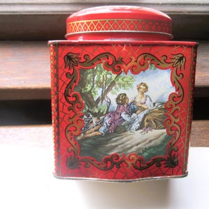 May include: A vintage red tin with a lid, featuring a decorative scene of people in a garden. The tin has gold accents and a geometric pattern. The lid also has a gold geometric design.