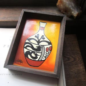 May include: A framed painting of a stylized vase with black and white geometric designs on a red and orange background. The vase is on a yellow background.