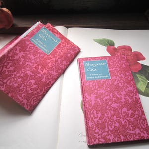 May include: Two copies of the book "Bhagavad-Gita: A Book of Hindu Scriptures" with pink floral patterned covers. One book is open, and the other is closed. The books are on a white surface with a floral illustration.