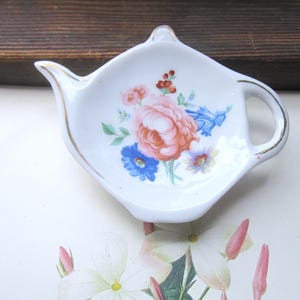 May include: A white ceramic tea bag holder shaped like a miniature teapot, featuring a central floral bouquet with a large pink rose, blue flowers, and green leaves. The rim and handle are accented with a thin gold trim, creating a delicate appearance.