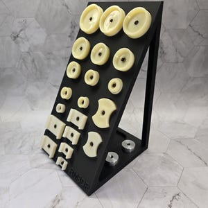 May include: A black angled display stand featuring an array of cream-colored circular, square, and curved components with central metal inserts. The base of the stand holds two silver circular discs, and the text RM3DP is embossed on the lower front.